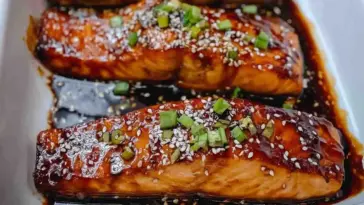 Baked teriyaki salmon fillets glazed with a rich sweet-savory sauce, topped with sesame seeds and chopped green onions, served in a baking dish.