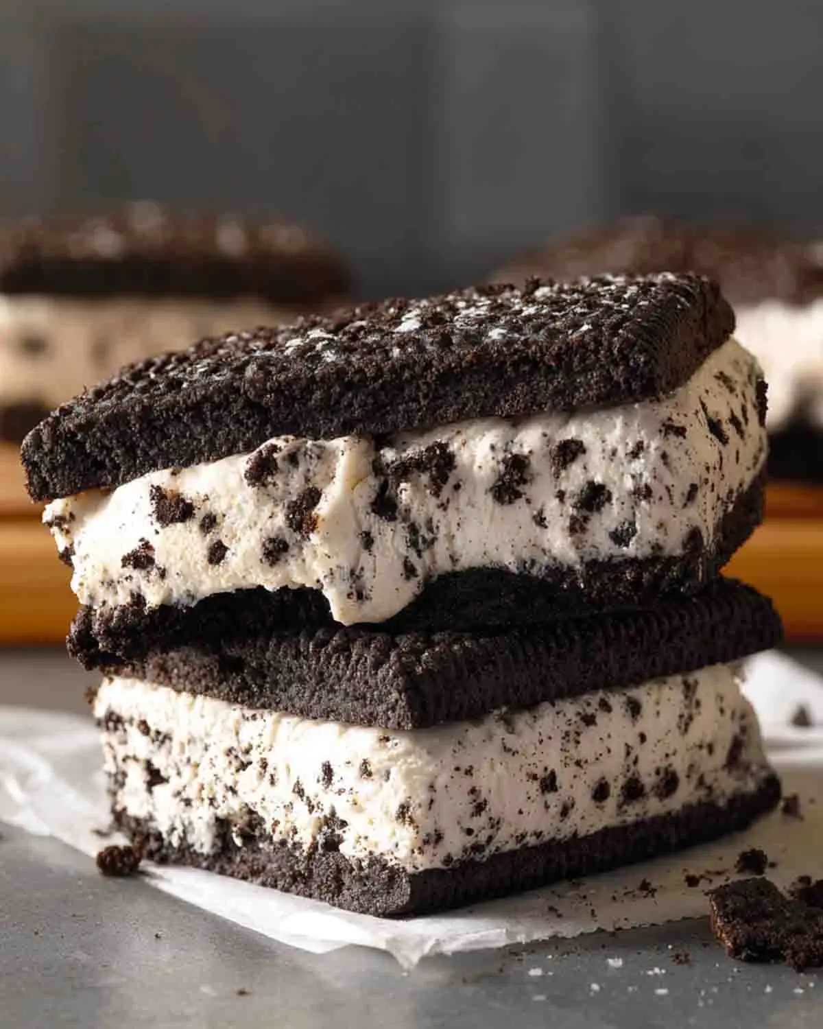 Stacked Oreo ice cream sandwiches with thick cookies and creamy cookies-and-cream filling, slightly melting with visible cookie crumbs on parchment paper.