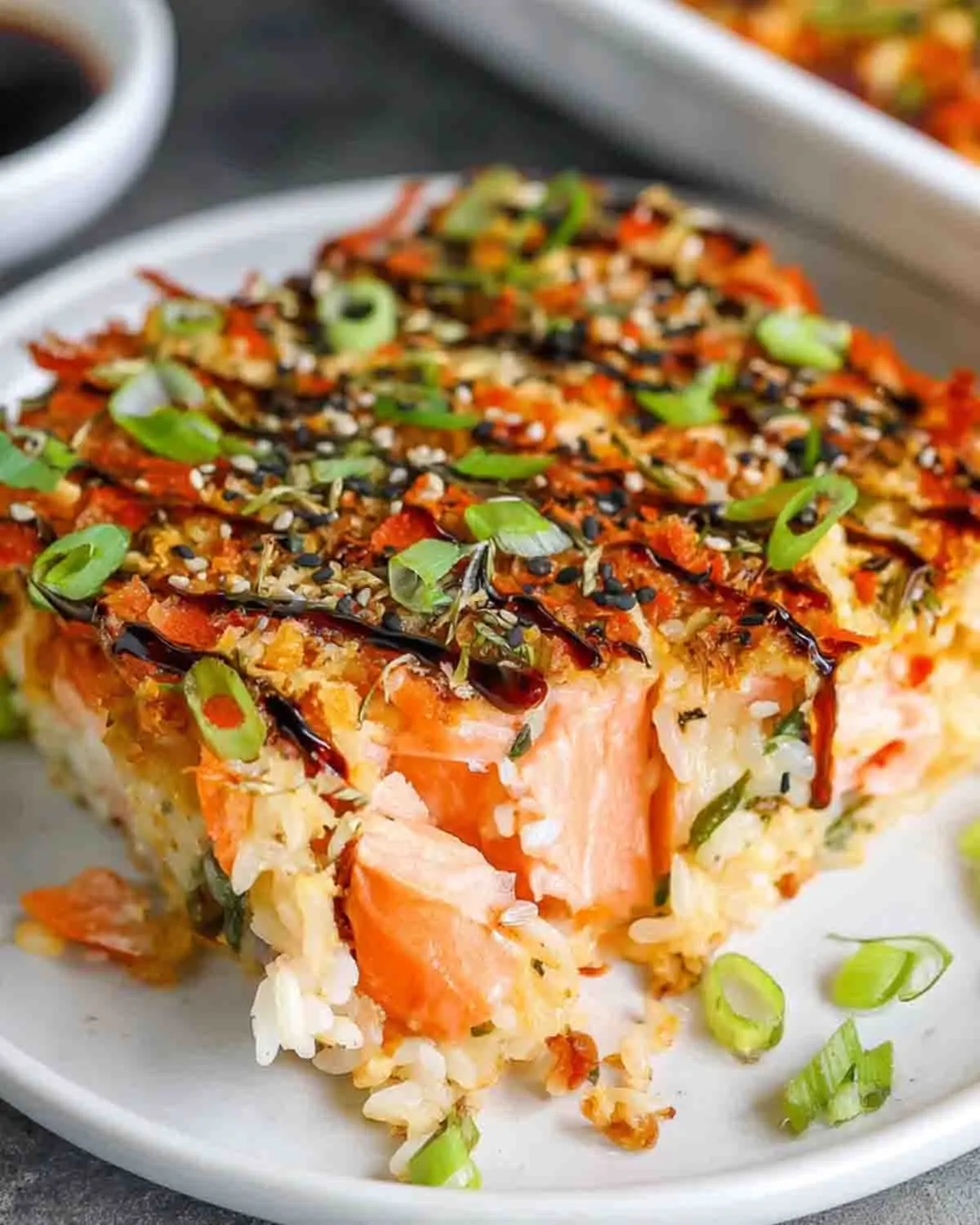 Close-up slice of salmon sushi bake served on a plate, featuring layered sushi rice, tender salmon chunks, drizzled sauce, sesame seeds, and fresh green onions on top