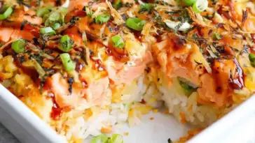 Close-up of creamy salmon sushi bake with fluffy rice layers, drizzled spicy mayo, furikake seasoning, and fresh green onions in a baking dish