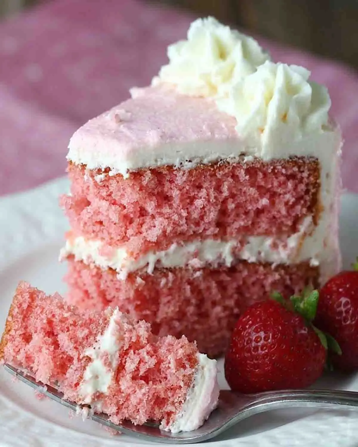 Close-up of a moist pink strawberry layer cake with creamy white frosting, a soft fluffy crumb, and fresh strawberries on the side, served on a plate with a fork.