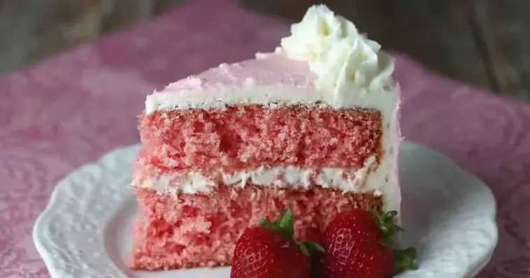 Moist strawberry layer cake slice with pink crumb and creamy white frosting, topped with whipped cream and served with fresh strawberries on a plate.