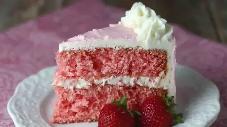 Moist strawberry layer cake slice with pink crumb and creamy white frosting, topped with whipped cream and served with fresh strawberries on a plate.